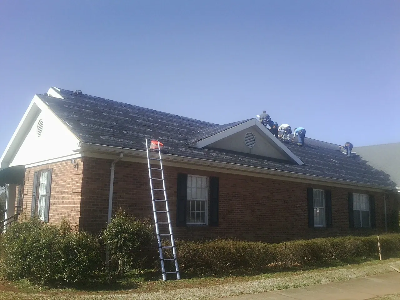 Crew tearing shingles off a brick ranch roof with a ladder set — Emergency Roof Repair work by our crew in Pilger