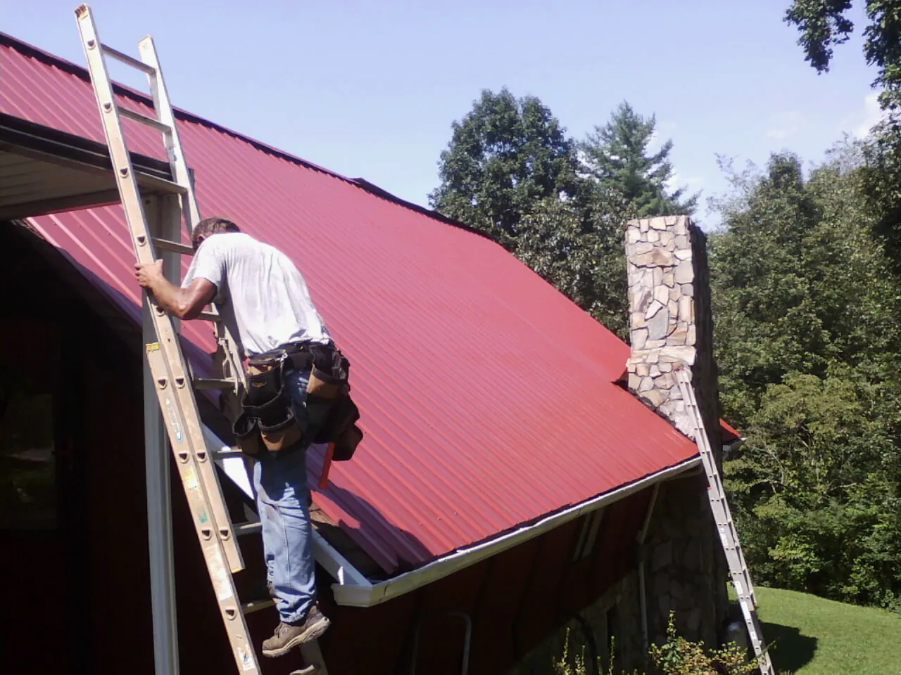 Roofer on a ladder beside a red metal roof and stone chimney — Soffit & Fascia Repair by Roofing Brothers in Pilger